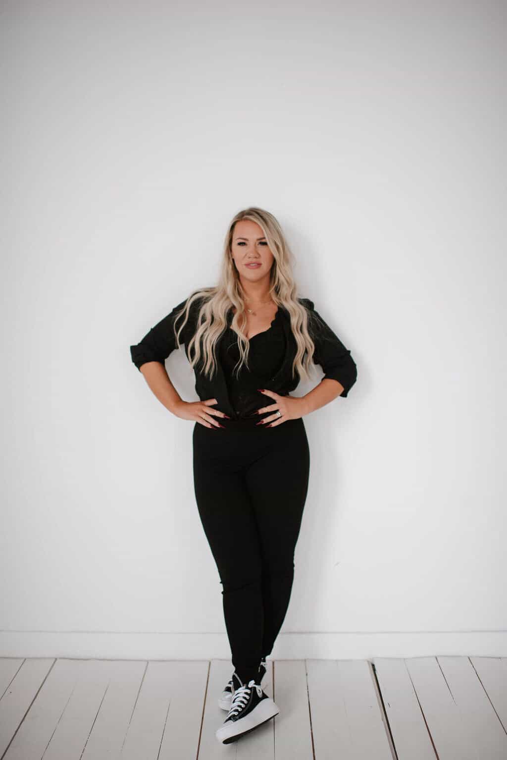 Woman standing confidently with hands on hips against a white background, dressed in black attire with contrasting white sneakers.