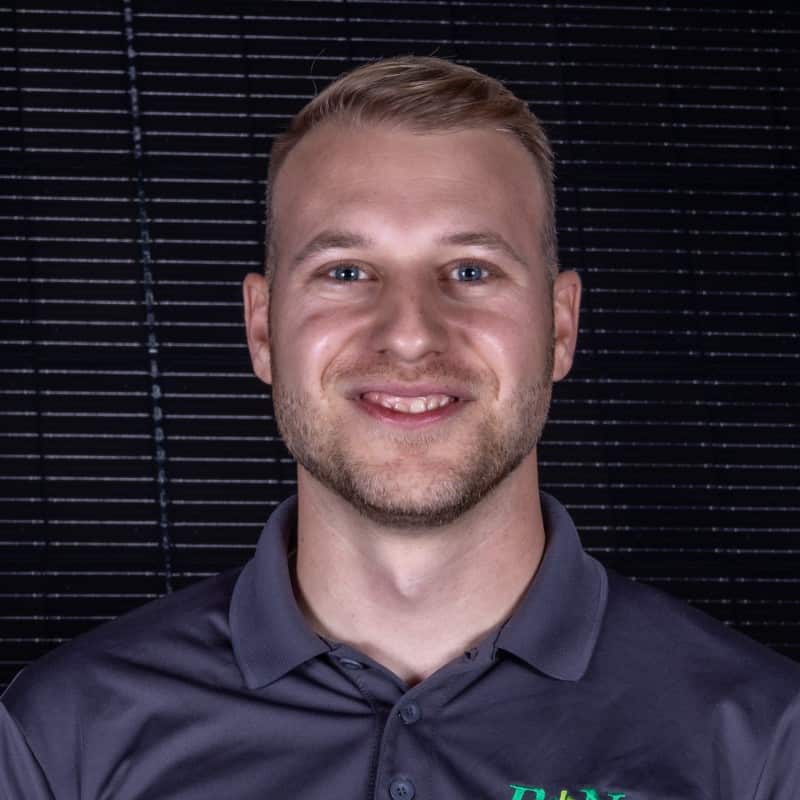 A smiling man with a short haircut, wearing a collared shirt with a logo on it, posed in front of a dark, vertical blind background.