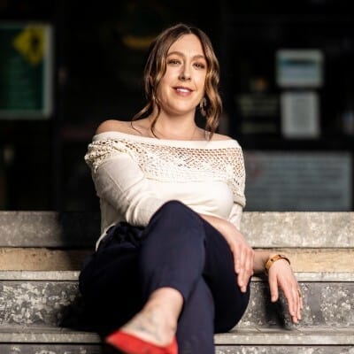 A person sitting with crossed legs on a set of stone steps, smiling at the camera, exuding a relaxed and confident vibe in a stylish outfit.