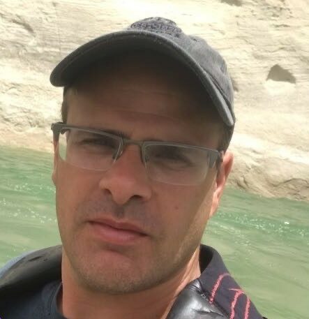 Man wearing glasses and a cap posing in front of a calm body of water with earthy cliffs in the background.