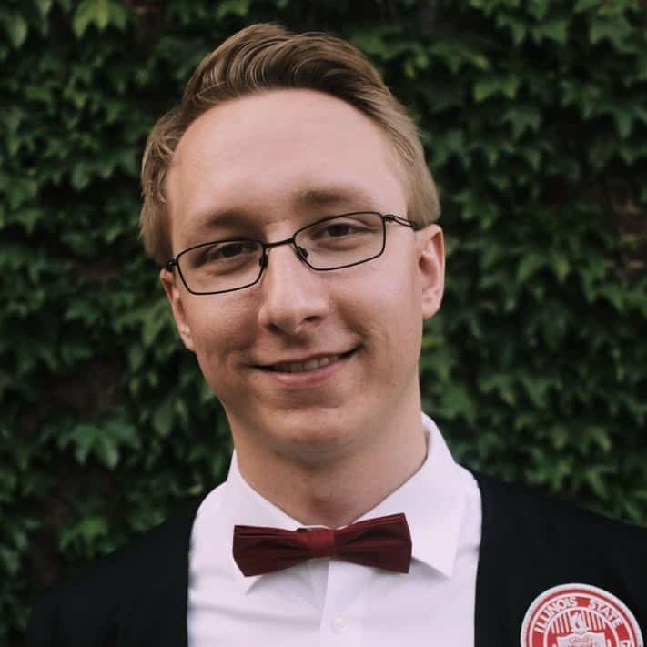 A man in a white shirt and bow tie smiling gently with a lush green hedge in the background.