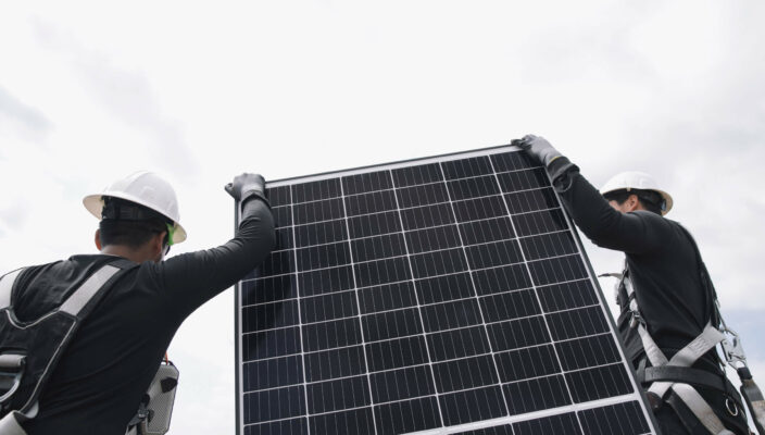 Workers in hard hats and safety gear installing a solar panel under a cloudy sky.