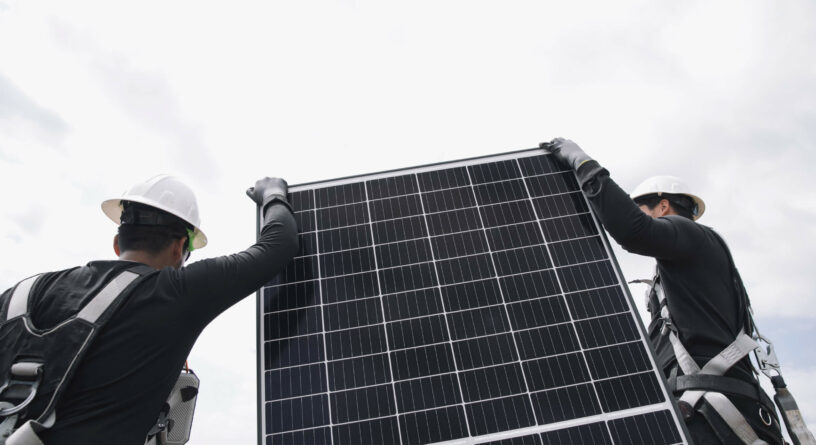 Workers in hard hats and safety gear installing a solar panel under a cloudy sky.