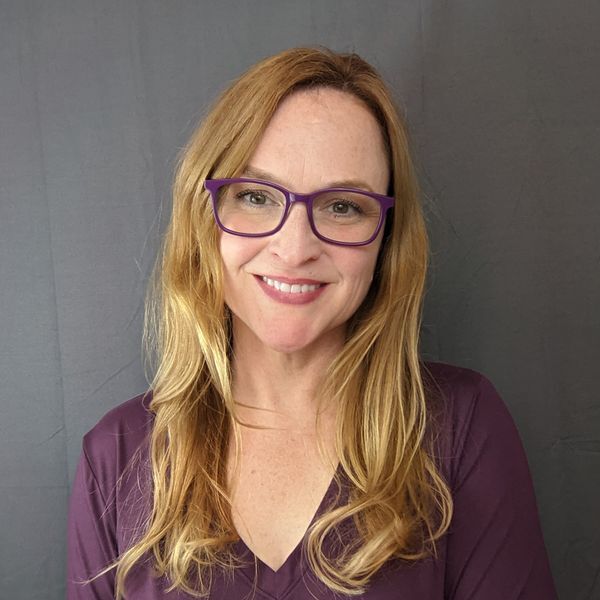 A smiling woman with long wavy blond hair wearing purple-framed glasses and a coordinated purple blouse against a grey background.