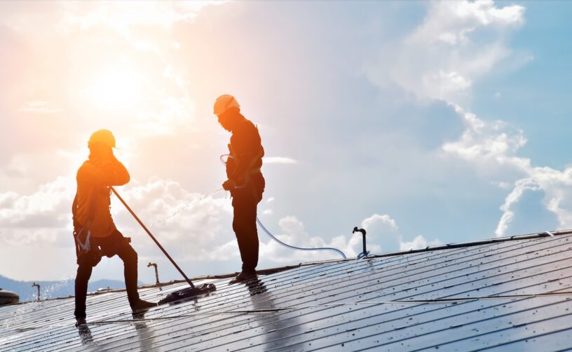 Two workers installing solar panels on a rooftop under a bright sunny sky.