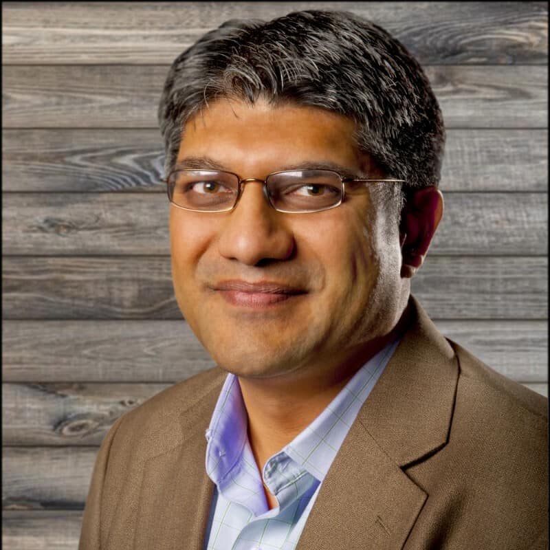 A professional portrait of a smiling man with glasses, wearing a blazer over a striped shirt, against a rustic wooden background.