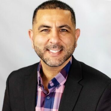 A professional headshot of a smiling man wearing a plaid shirt and jacket.