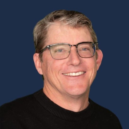 A smiling man wearing glasses with a blue background.