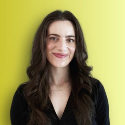 A smiling woman with long brown hair, wearing a black top, against a bright yellow background.