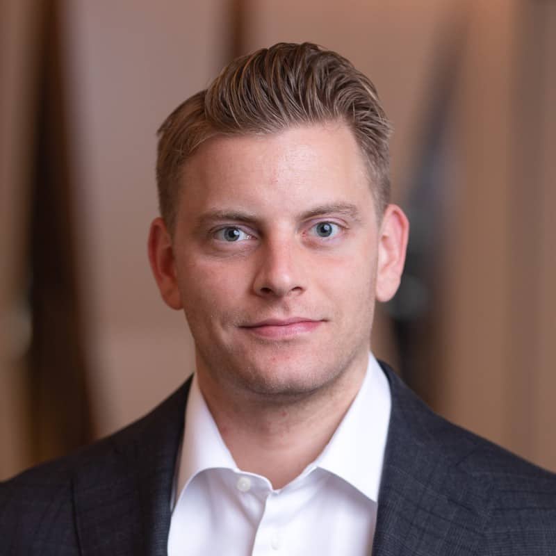 A professional headshot of a man with slicked-back hair, wearing a suit with an open-collar white shirt, set against a blurred indoor background.