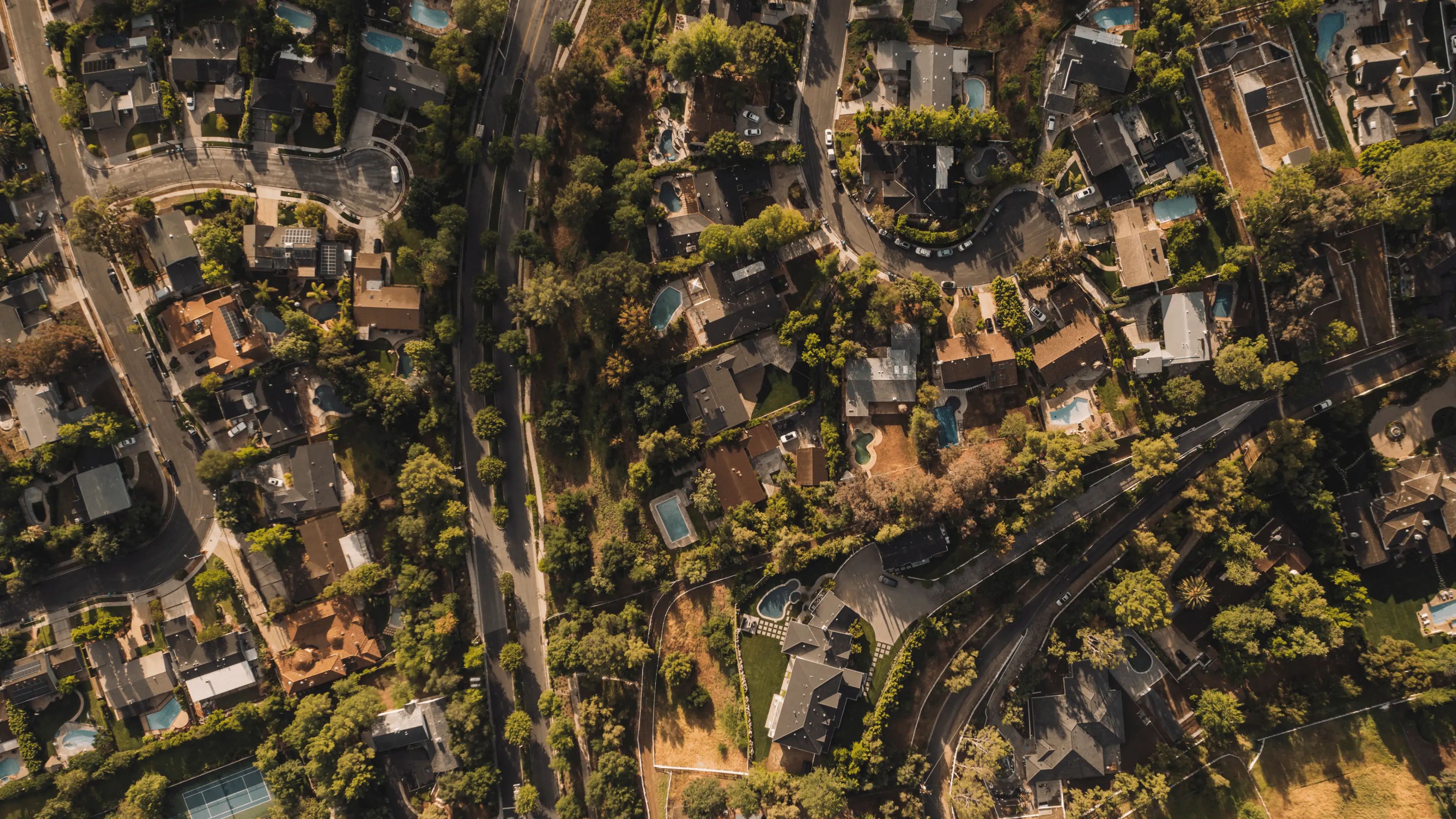 Aerial view of a residential area showing a tapestry of houses, pools, and winding streets amidst a canopy of trees.