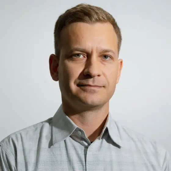 A professional headshot of a man with a subtle smile, wearing a light blue button-up shirt against a neutral background.