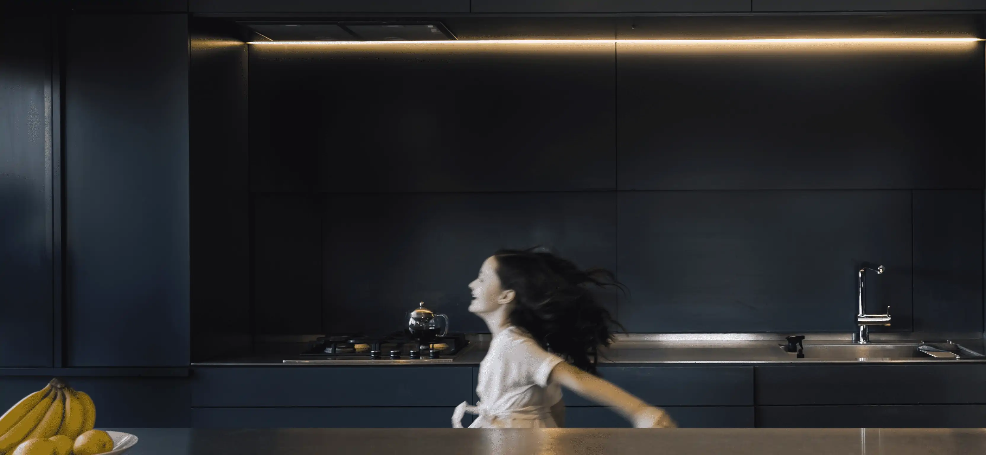 A blurred young girl in motion, adding a sense of liveliness and action to the sleek, modern kitchen with its dark cabinets and ambient lighting.