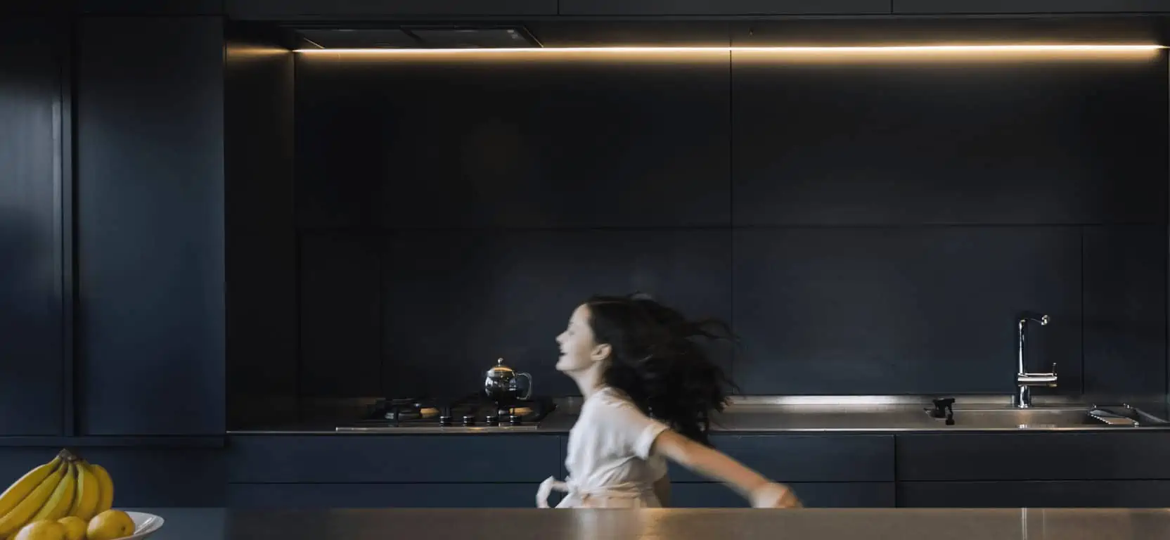 A young girl in a white shirt passing by a sleek modern kitchen with motion blur, indicating movement and life within a contemporary home setting.