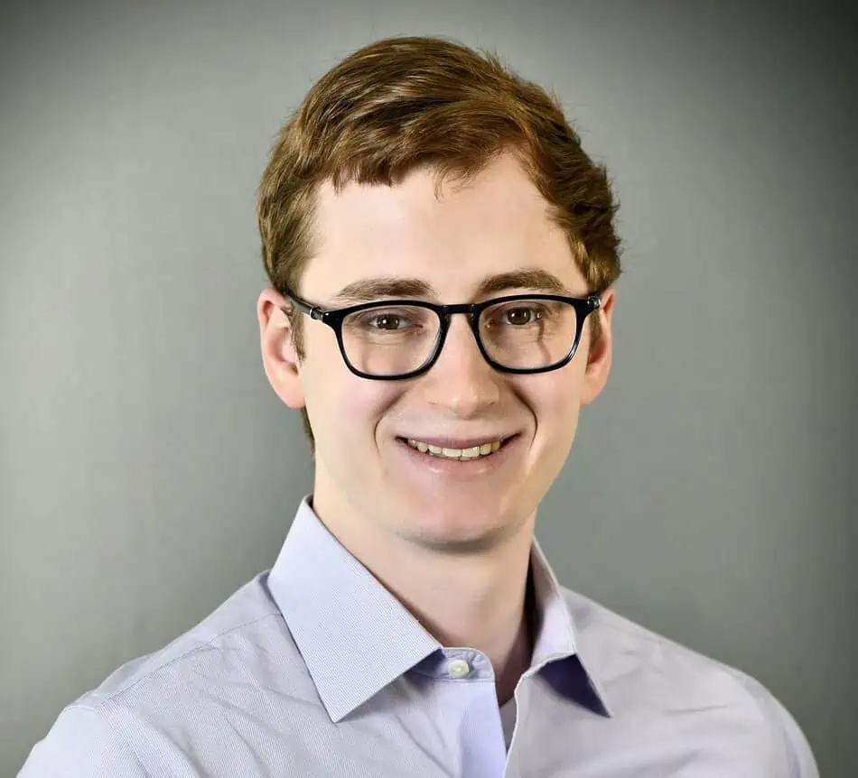 A smiling individual with glasses wearing a light blue button-up shirt against a gray backdrop.