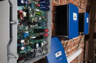 An open industrial electrical panel revealing intricate electronic components mounted on a brick wall accompanied by two closed blue electrical boxes