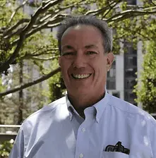 A smiling man outdoors with trees and a building in the background.