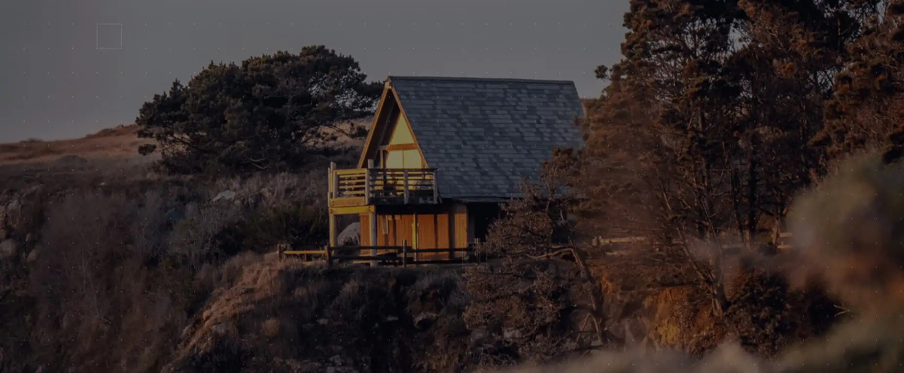 A solitary cabin perched on a cliffside, basking in the warm glow of interior lights against the dusky twilight.