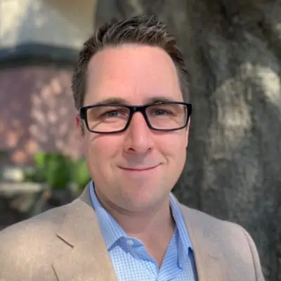 A smiling man with glasses, wearing a blazer and a blue checkered shirt, with a softly blurred background emphasizing his cheerful expression.